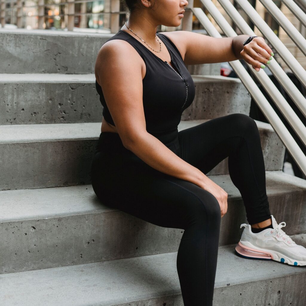 Woman in black sports clothing sitting on outdoor stairs, checking wristwatch.